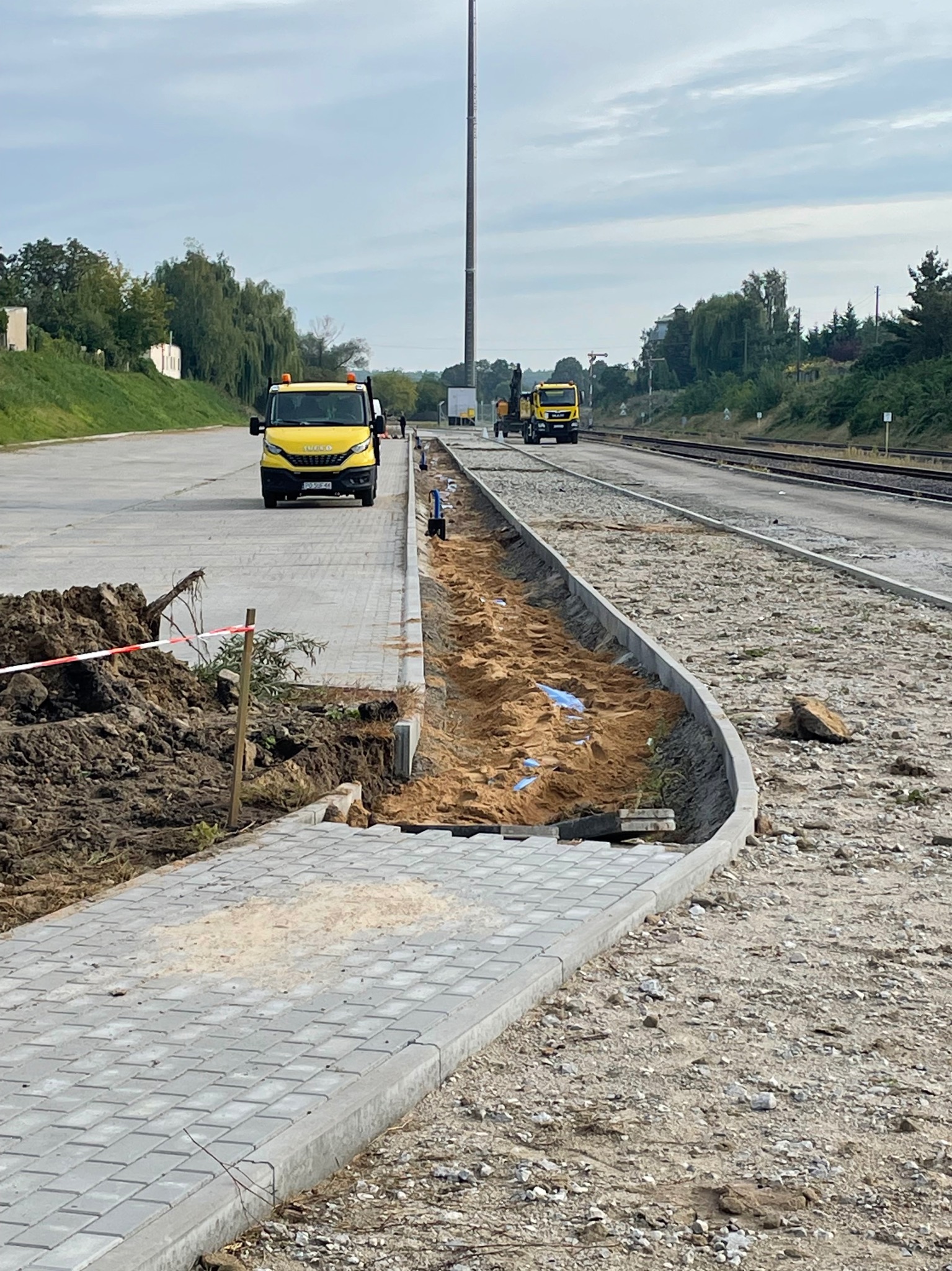 Fragment układanej szarej kostki brukowej, widoczne żółte samochody dostawcze na tle torów kolejowych i ziemi. Prace w toku, widoczne materiały budowlane.