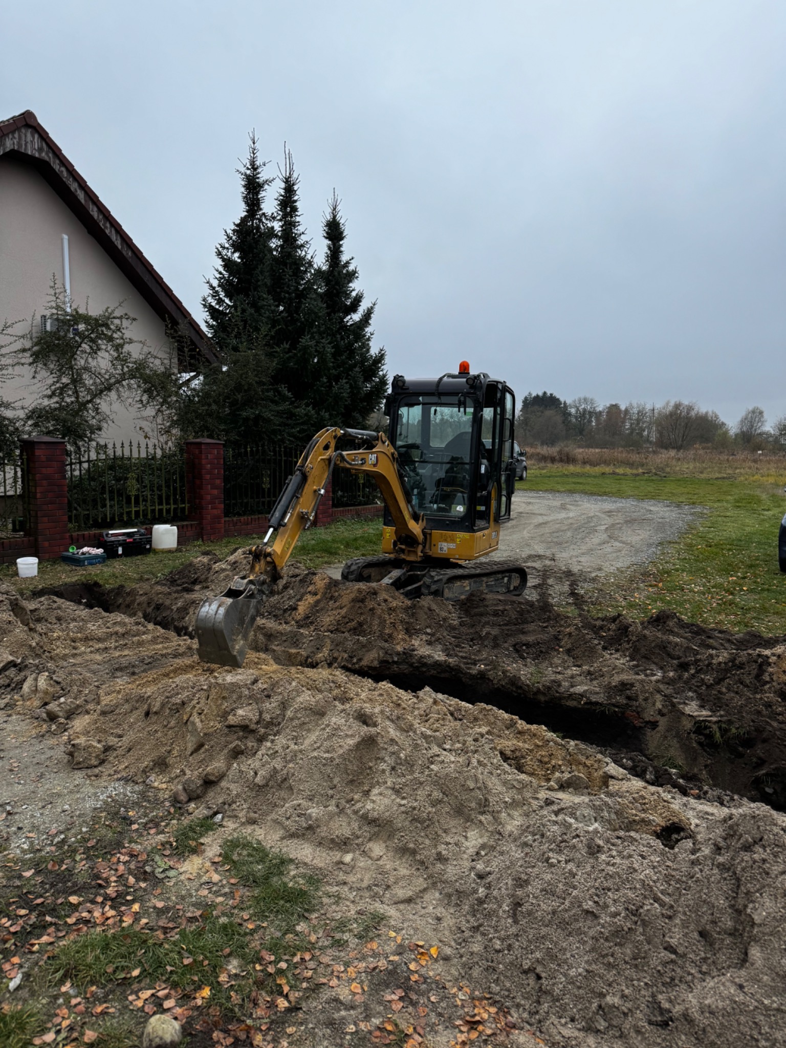 Żółta minikoparka CAT podczas wykopu rowu na działce. W tle dom z czerwonej cegły i zielone drzewa. Pochmurne niebo.