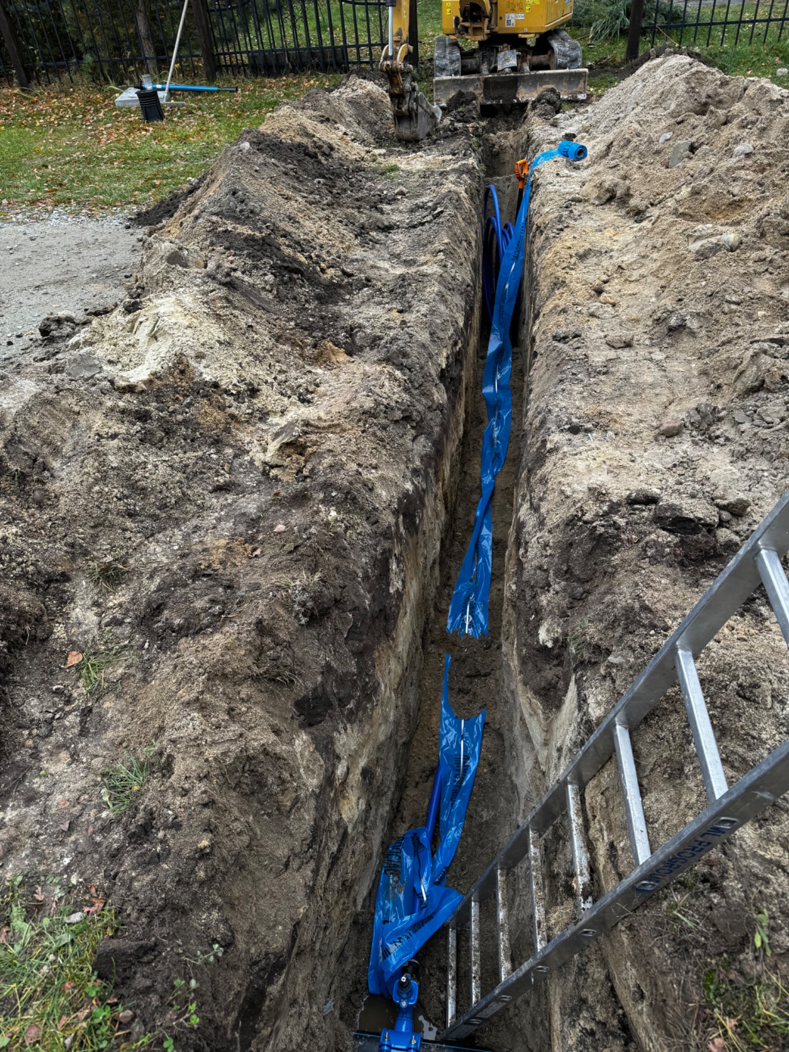 Wykop z rurami w niebieskiej folii ochronnej, widoczna drabina i koparka w tle. Instalacja w ziemi.
