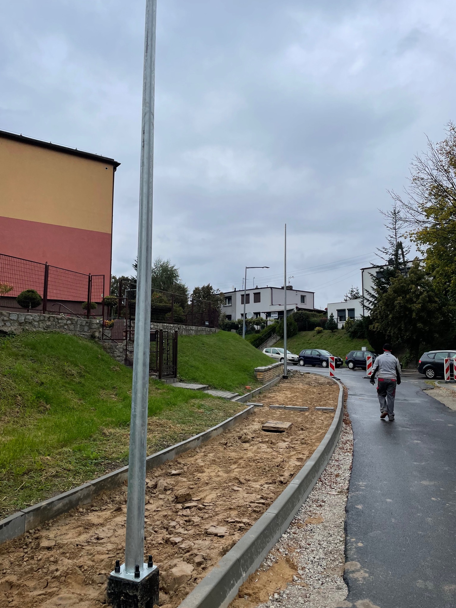 Przygotowanie podłoża pod chodnik z obrzeżami, widoczny piasek i fragment drogi asfaltowej. W tle budynki mieszkalne i słup oświetleniowy.