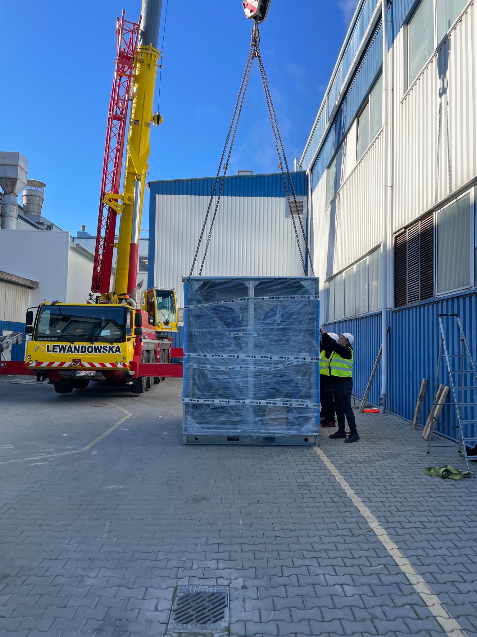 Montaż agregatu chłodniczego dźwigiem na zewnątrz budynku przemysłowego, pracownicy w kaskach i kamizelkach odblaskowych nadzorują proces.