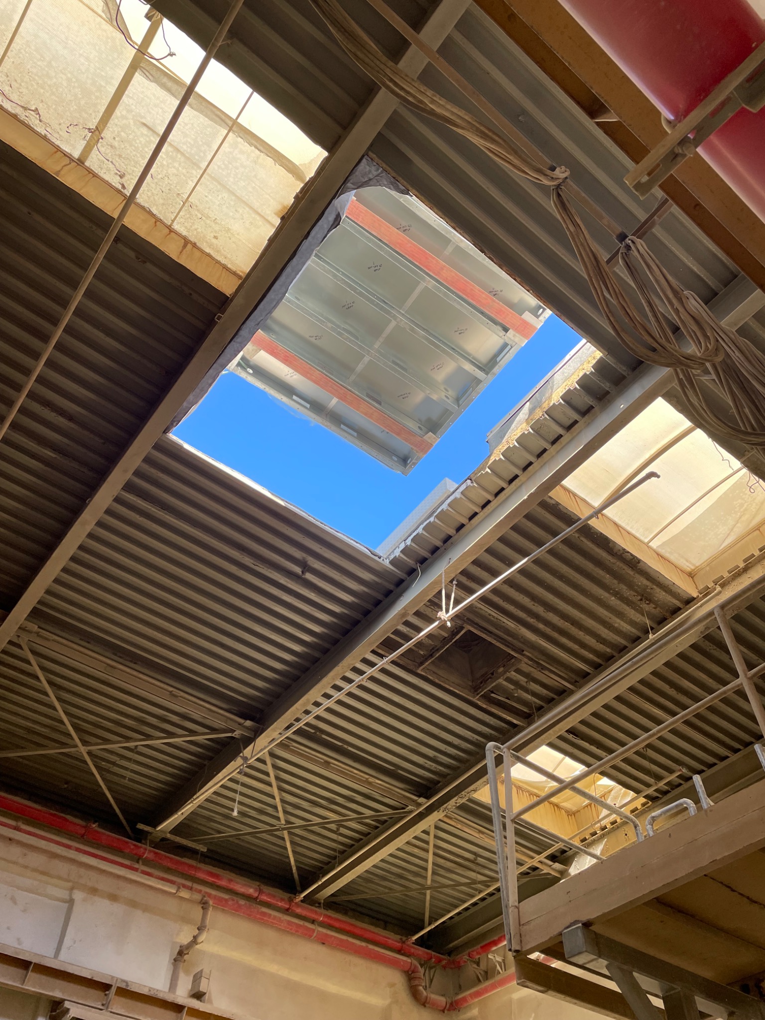 Metalowa konstrukcja stropu z otworami, przez które widać błękitne niebo. Widoczne kable, rury i elementy konstrukcyjne. Ujęcie z dołu, surowy industrialny styl.