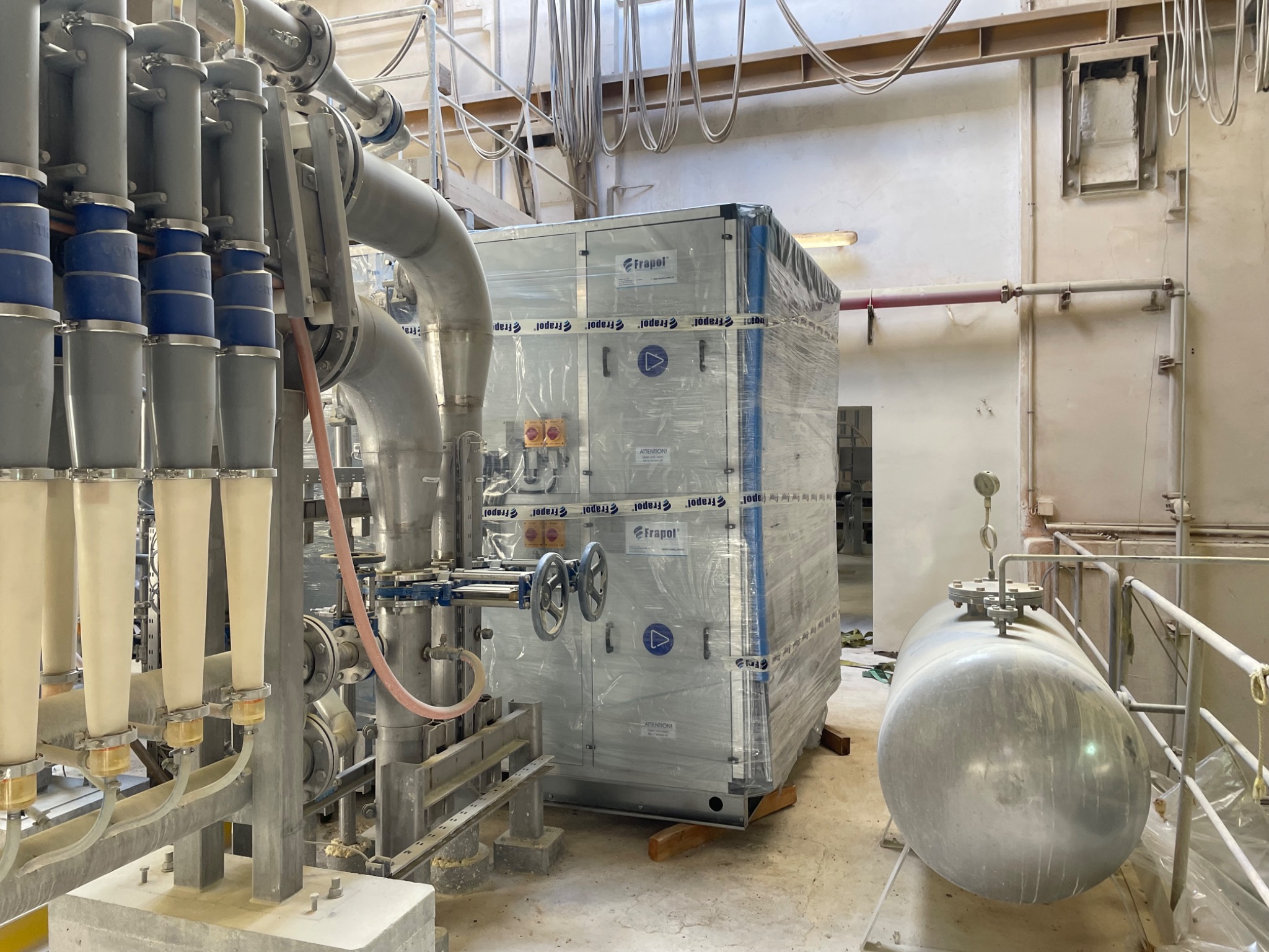 Nowa, owinięta folią przemysłowa centrala wentylacyjna Frapol na drewnianych podkładkach w pomieszczeniu technicznym z rurami i zbiornikiem ciśnieniowym.