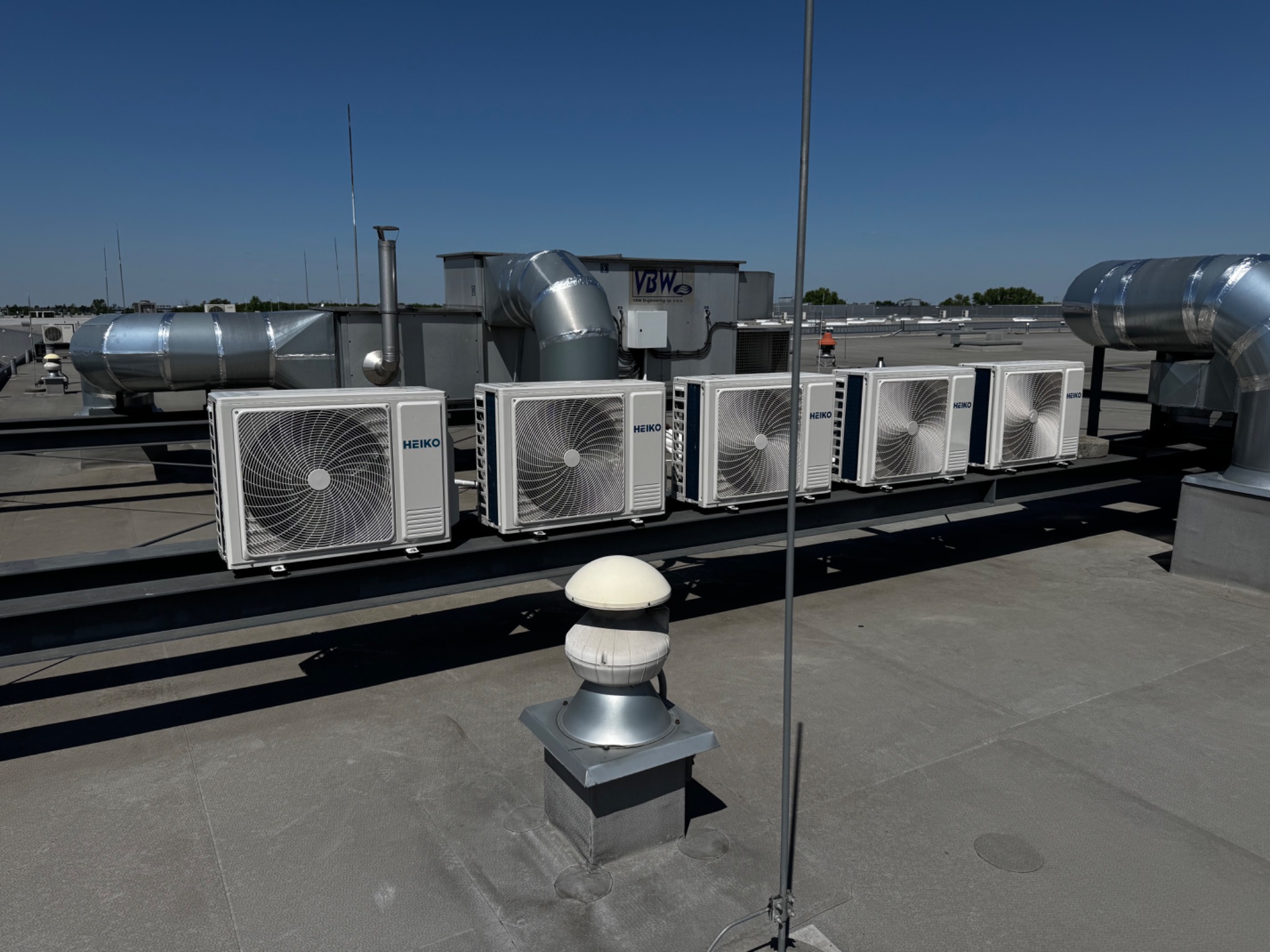 Rząd jednostek zewnętrznych klimatyzacji HEIKO na dachu budynku, połączonych metalowymi rurami wentylacyjnymi, w tle niebieskie niebo. Widok z góry.