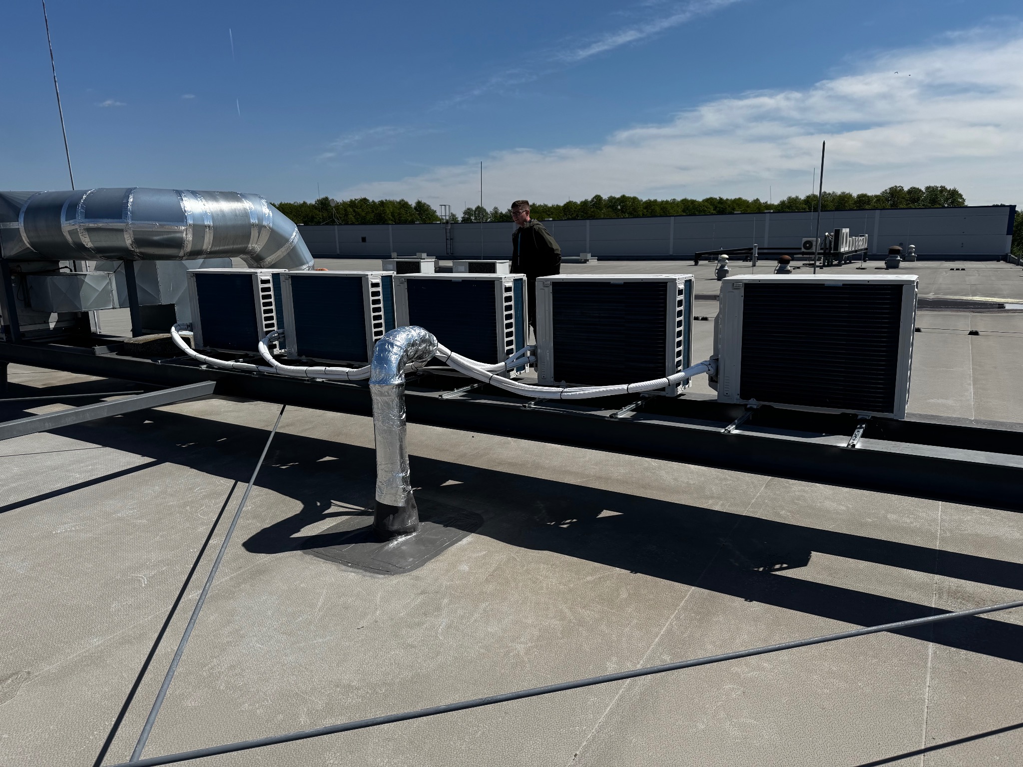 Rząd klimatyzatorów na dachu budynku, połączonych srebrnymi rurami. Na tle błękitne niebo i zieleń drzew. Widoczny fragment instalacji wentylacyjnej.