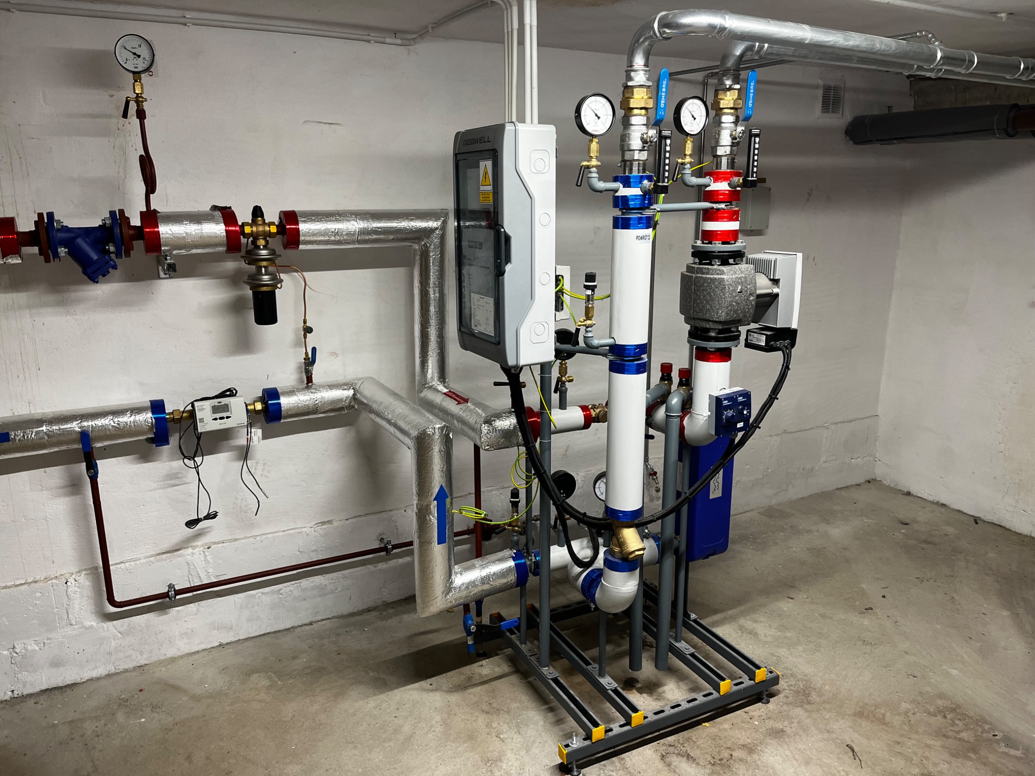 Złożona instalacja grzewcza z rurami, zaworami i manometrami, umieszczona na metalowej ramie w pomieszczeniu z betonową podłogą i białymi ścianami.