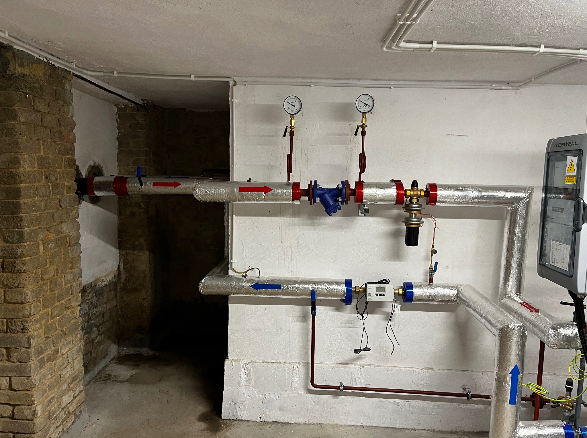 Izolowane rury z zaworami i manometrami w piwnicy. Widoczne elementy instalacji grzewczej oraz fragment ściany z cegły. Strzałki wskazują kierunek przepływu.