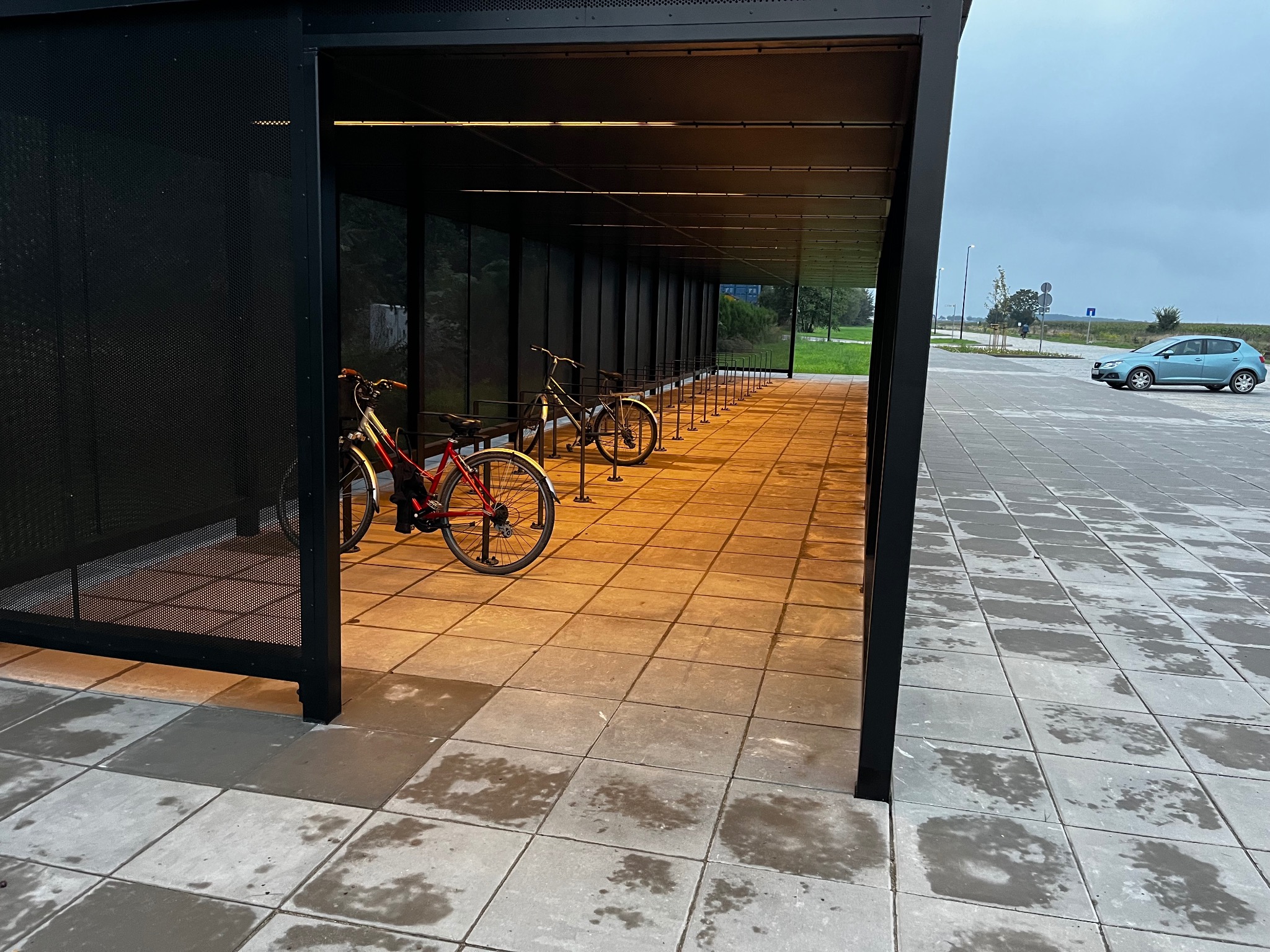 Zadaszony parking rowerowy z kostki brukowej, oświetlony od wewnątrz, z zaparkowanymi rowerami. W tle widoczny parking i samochód.