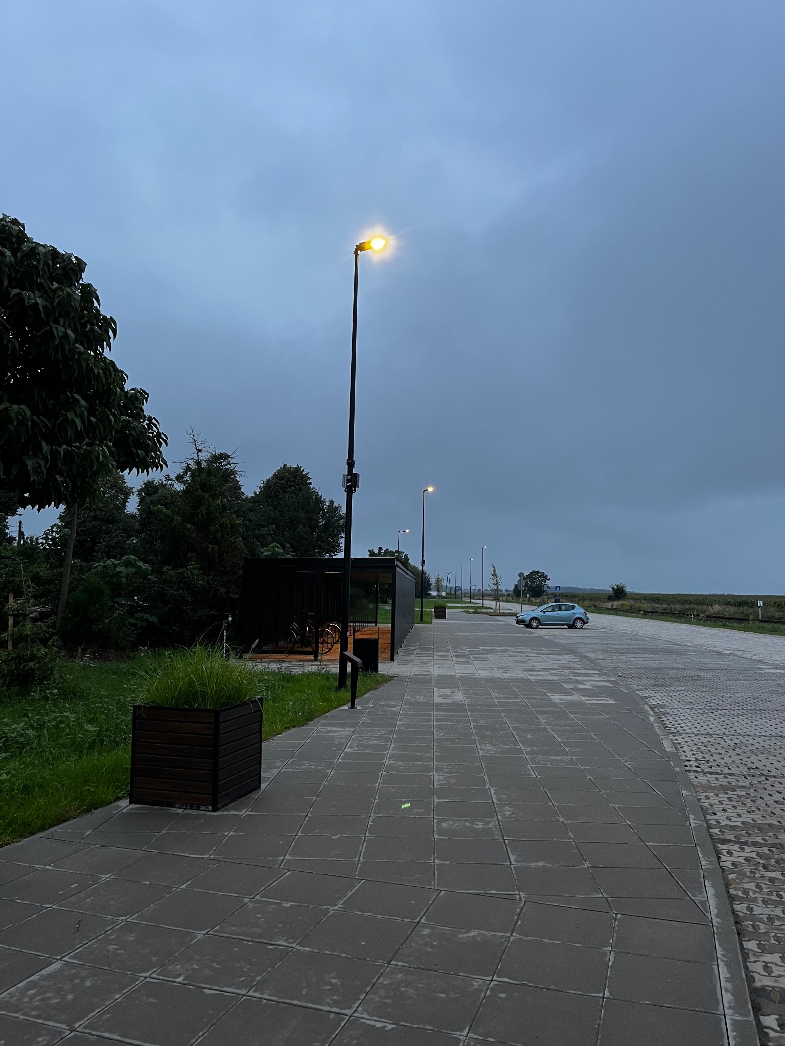 Szara kostka brukowa wzdłuż drogi z lampami ulicznymi i błękitnym autem w tle, oświetlona wieczornym światłem. Widoczny przystanek rowerowy i zieleń.