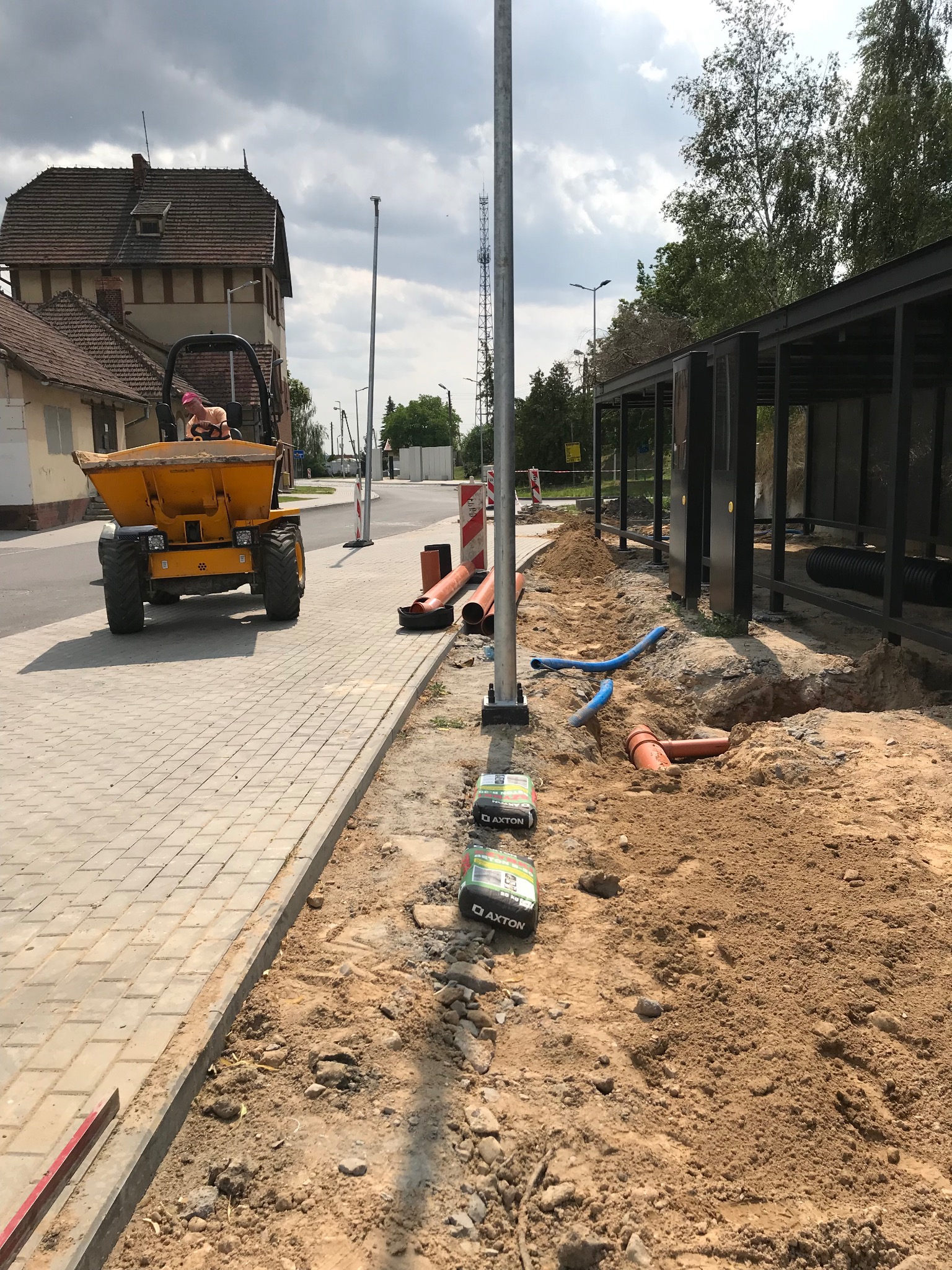 Żółta wywrotka na chodniku z kostki brukowej obok wykopu z rurami i workami cementu Axton. W tle budynek i konstrukcja przystanku.