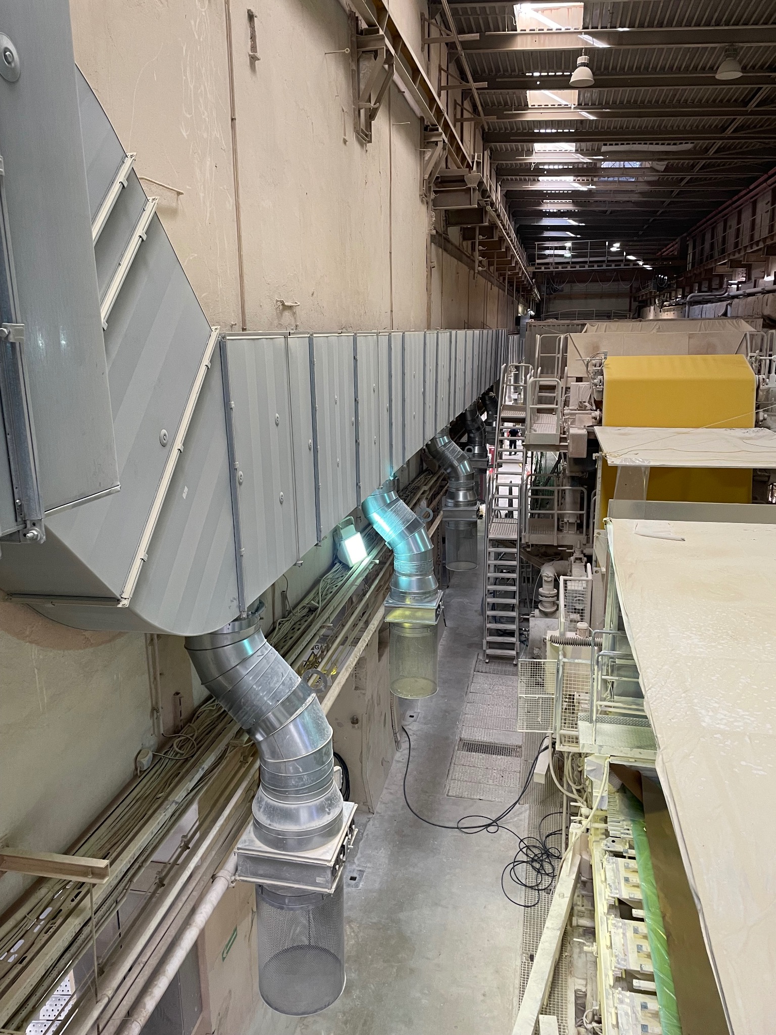 Wysoka hala przemysłowa z systemem wentylacji kanałowej pod sufitem i maszynami produkcyjnymi w tle, widok z góry w neutralnej tonacji kolorystycznej.