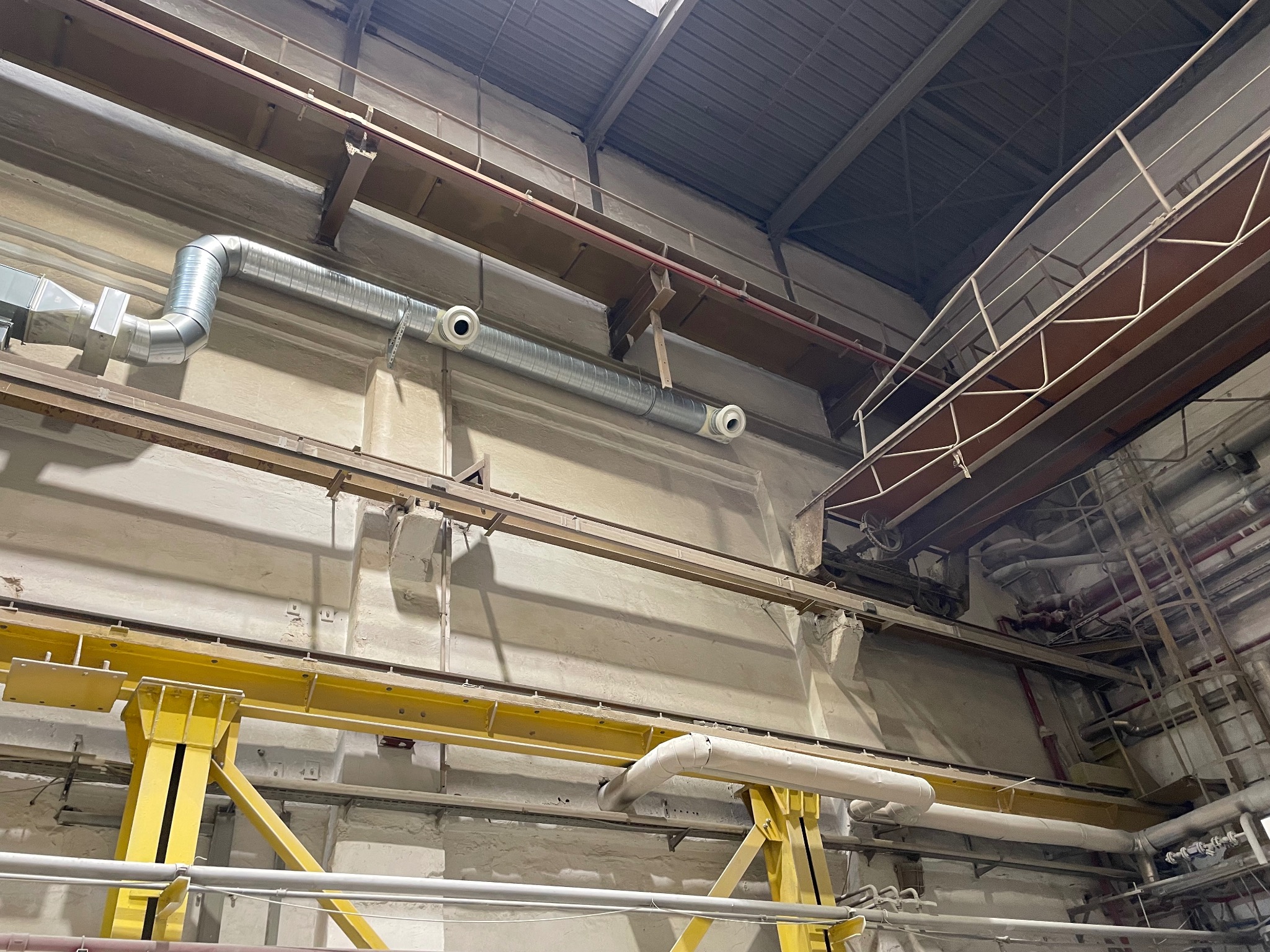 Wnętrze hali przemysłowej z widocznymi rurami wentylacyjnymi i elementami konstrukcyjnymi pod sufitem. Metalowe rury i żółta konstrukcja stalowa.