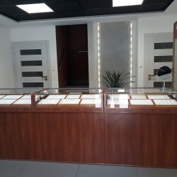 A&J Jubiler Justyna Zaremba - Wnętrze sklepu jubilerskiego z drewnianymi witrynami wypełnionymi biżuterią, obok witryny regał z biżuterią i figurką gnoma na górze, w tle drzwi z przeszkleniami i fragment ceglanej ściany.