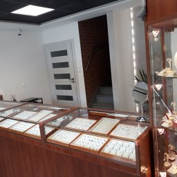 A&J Jubiler Justyna Zaremba - Wnętrze sklepu jubilerskiego z przeszklonymi gablotami wypełnionymi biżuterią, widoczne naszyjniki, bransoletki i pierścionki na białych ekspozytorach, w tle schody i drzwi.