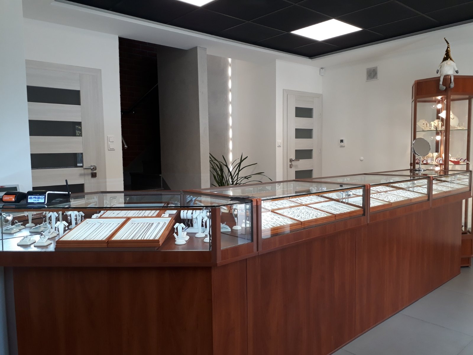 Wnętrze sklepu jubilerskiego: długi, drewniany lada z przeszklonymi gablotami prezentującymi srebrną biżuterię, w tle witryna z oświetleniem i dekoracyjna figurka. Widoczny fragment schodów.