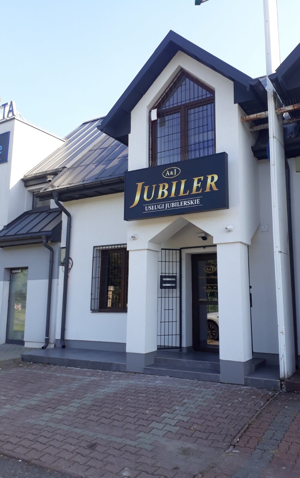 Fasada budynku z szyldem 'A&J Jubiler Usługi Jubilerskie' w złotych literach na czarnym tle, widok z zewnątrz na wejście z metalową kratą i okno z kratami, białe ściany, ciemny dach i kostka...