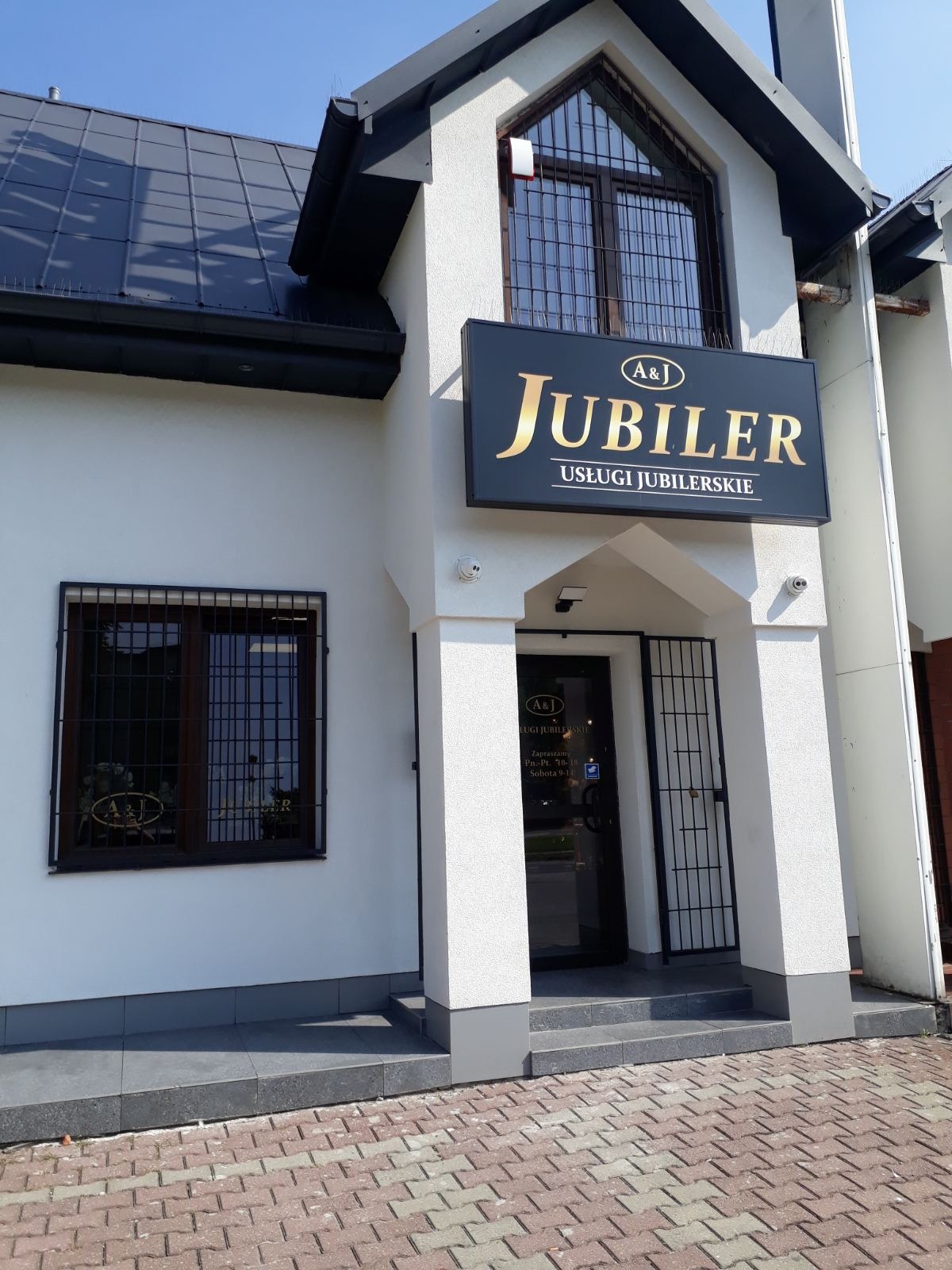 Fasada budynku z szyldem 'Jubiler A&J Usługi Jubilerskie' nad wejściem, okna z kratami, biała elewacja, ceglana kostka brukowa na chodniku.