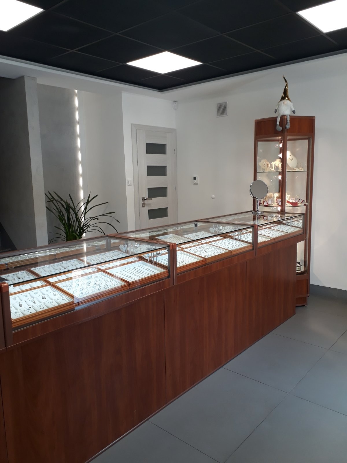 Wnętrze sklepu z biżuterią: długi drewniany lada z szklanymi gablotami wypełnionymi biżuterią, w tle witryna z ozdobami i figurka skrzata na górze.