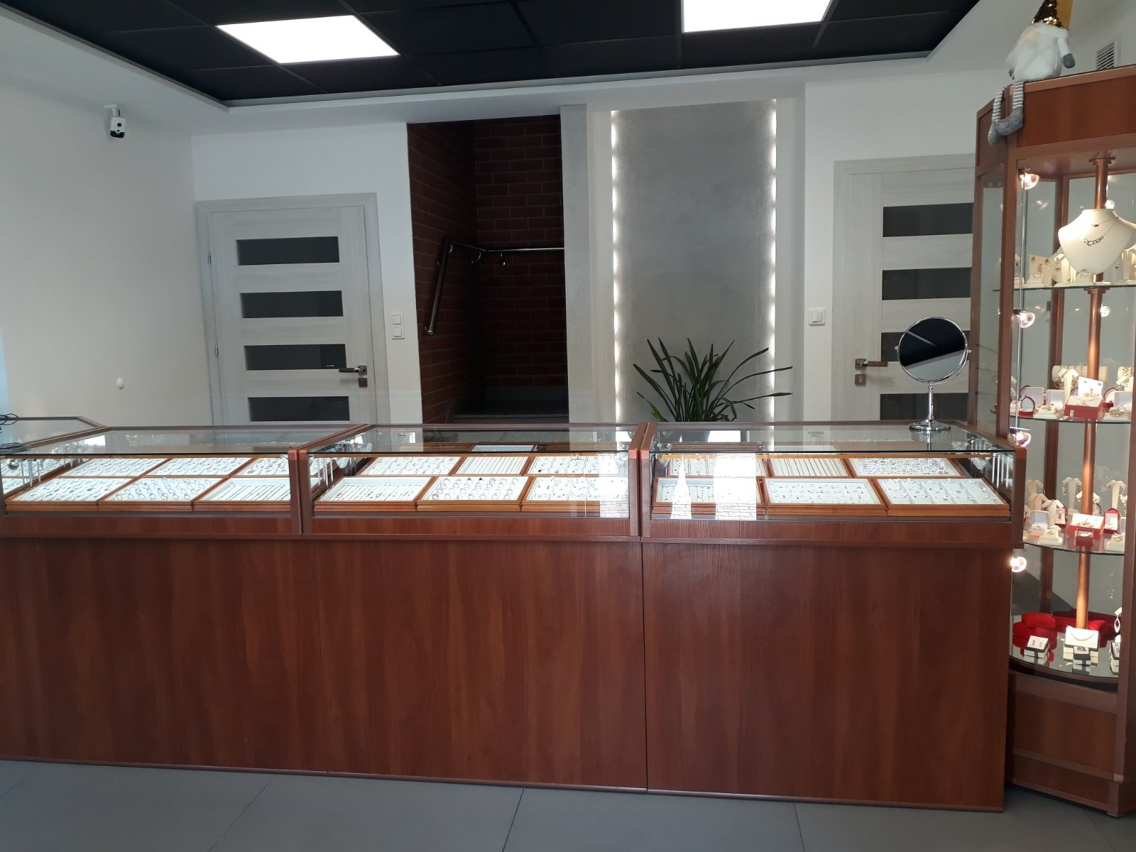 Wnętrze sklepu jubilerskiego z drewnianymi witrynami wypełnionymi biżuterią, obok witryny regał z biżuterią i figurką gnoma na górze, w tle drzwi z przeszkleniami i fragment ceglanej ściany.