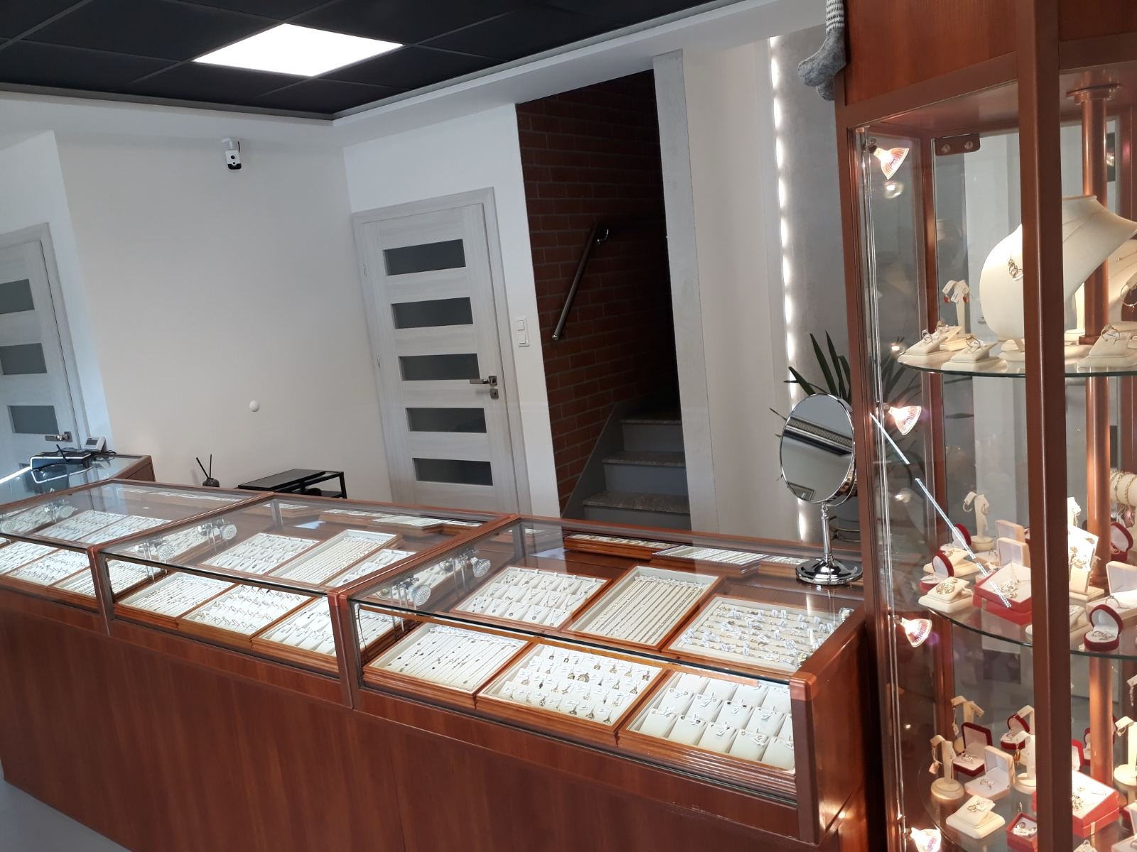 Wnętrze sklepu jubilerskiego z przeszklonymi gablotami wypełnionymi biżuterią, widoczne naszyjniki, bransoletki i pierścionki na białych ekspozytorach, w tle schody i drzwi.