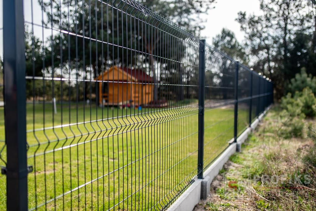 Nowoczesne, ciemne ogrodzenie panelowe z podmurówką betonową na trawniku, z widocznym drewnianym domkiem ogrodowym w tle.