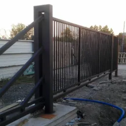 Czarna, przesuwna brama przemysłowa z pionowymi szczeblami, widoczna podczas montażu, z fragmentem ogrodzenia z drutem kolczastym w tle i zaparkowanym samochodem.