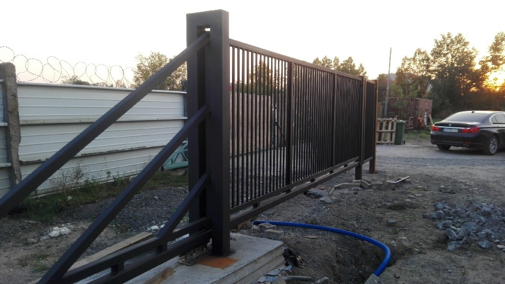 Czarna, przesuwna brama przemysłowa z pionowymi szczeblami, widoczna podczas montażu, z fragmentem ogrodzenia z drutem kolczastym w tle i zaparkowanym samochodem.