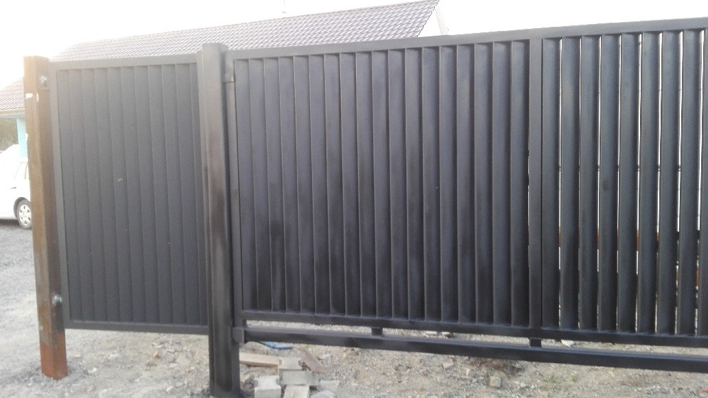 Nowoczesna, metalowa brama przesuwna w kolorze grafitowym z pionowymi panelami, widoczna z bliska na tle domu z jasną elewacją.