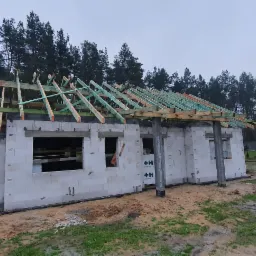 Surowa konstrukcja domu z białych bloczków, widoczne drewniane elementy konstrukcji dachowej, tło stanowią drzewa i pochmurne niebo.