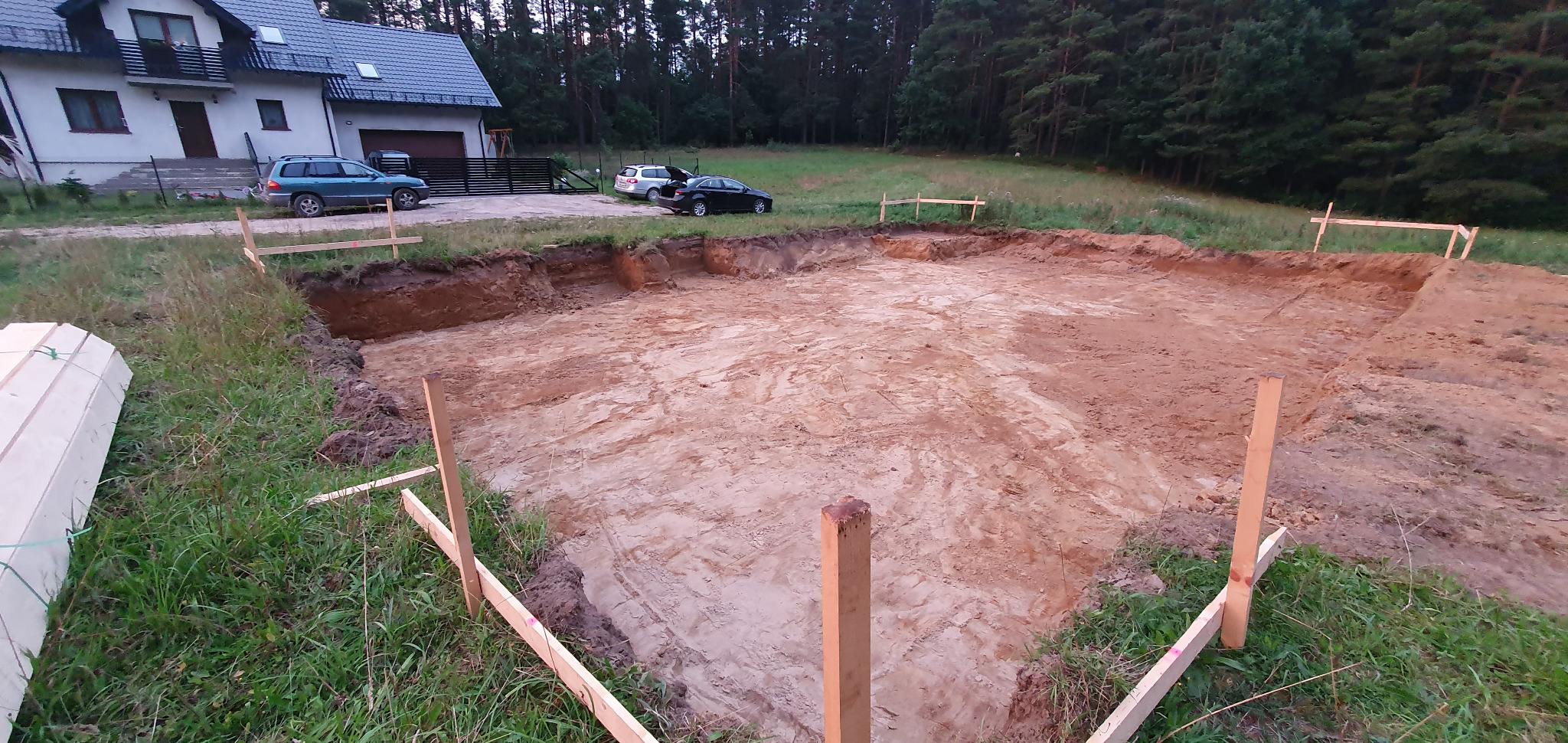 Szeroki wykop pod fundamenty budynku jednorodzinnego, widoczne drewniane słupki wyznaczające obrys, w tle dom z szarym dachem i las.