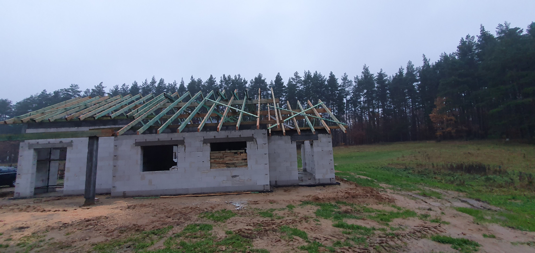 Budowa domu jednorodzinnego w stanie surowym otwartym z widoczną konstrukcją dachu na tle lasu i pochmurnego nieba. Ściany z betonu komórkowego.