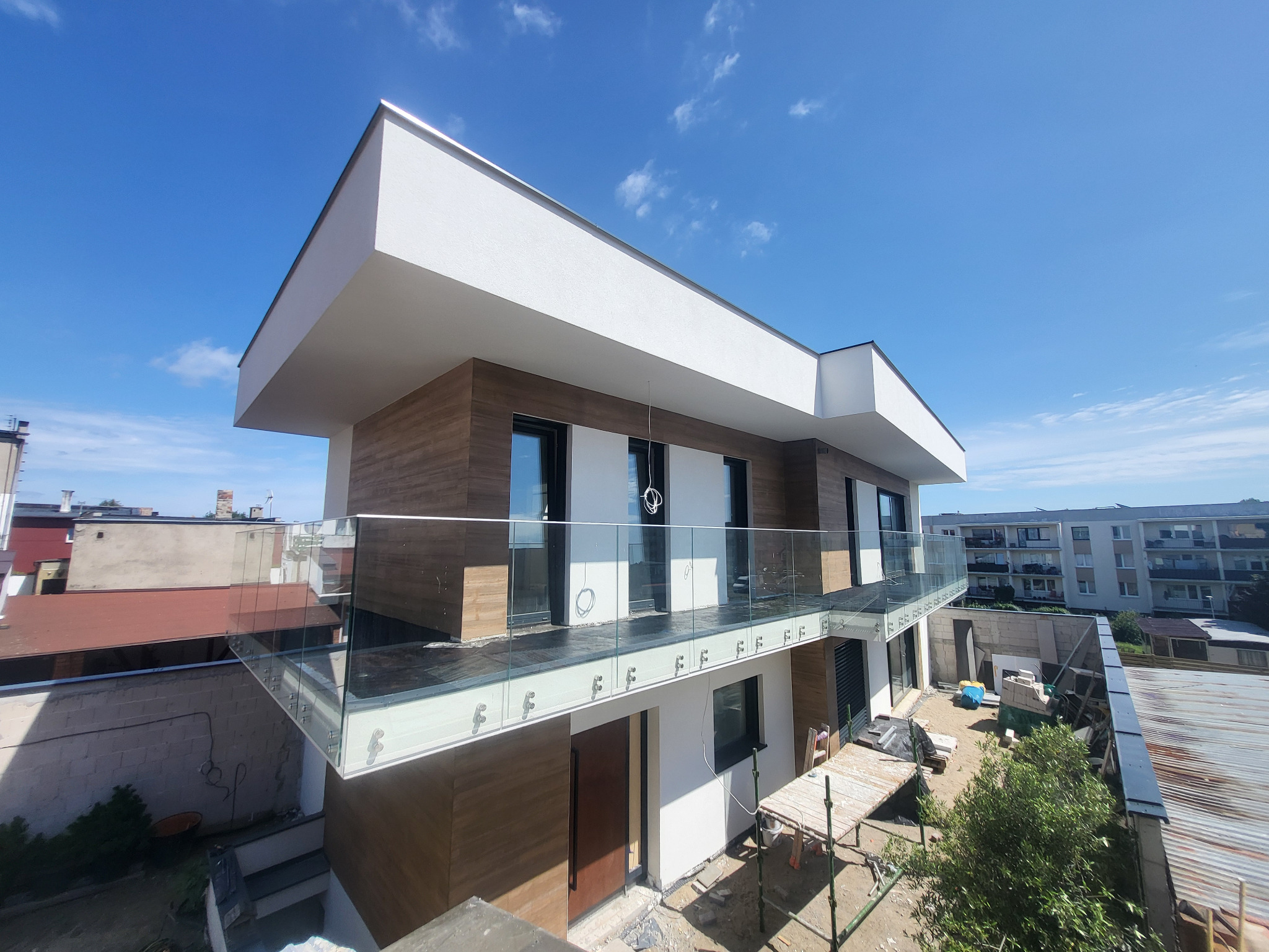 Nowoczesny, dwupiętrowy dom z balkonami z przezroczystymi balustradami, wykończony drewnopodobną okładziną i białym tynkiem, na tle błękitnego nieba.