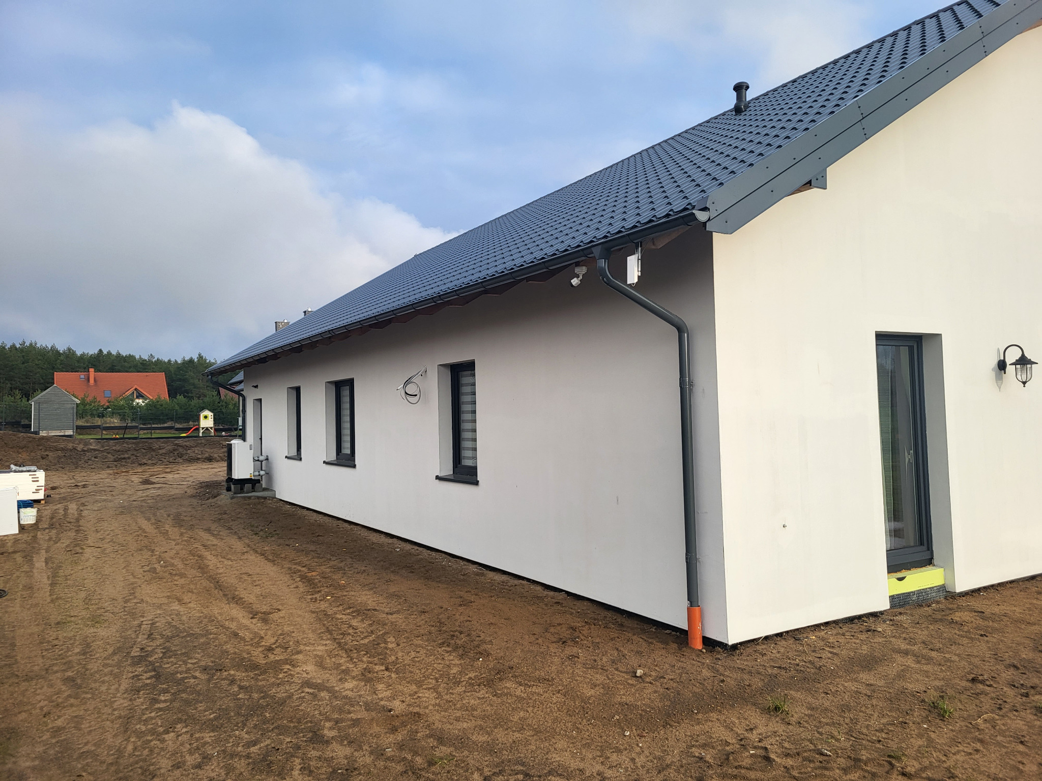 Nowoczesny, parterowy dom z szarą dachówką i białymi ścianami, widok z boku. Uwagę zwraca minimalistyczna lampa zewnętrzna i pomarańczowa rura spustowa.