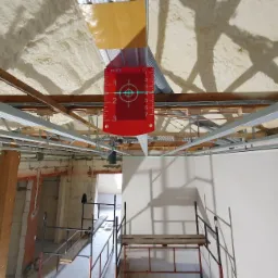 Montaż sufitu podwieszanego z widoczną izolacją piankową stropu i użyciem poziomicy laserowej, rusztowanie w tle.