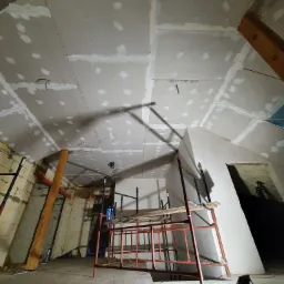 Poddasze w trakcie wykańczania: zamontowane płyty gipsowo-kartonowe na skośnym suficie, widoczne łączenia płyt wymagające szpachlowania, metalowe rusztowanie w centrum kadru, oświetlenie robocze...