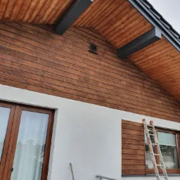 Wykończona drewniana elewacja domu z widocznymi oknami, fragmentem balkonu i drabiną opartą o ścianę, ukazująca detale drewnianej konstrukcji i połączenie z białą elewacją.