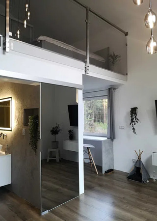 Nowoczesne wnętrze z antresolą, szklaną balustradą, lustrzanymi drzwiami do łazienki i drewnianą podłogą, oświetlone wiszącymi lampami.
