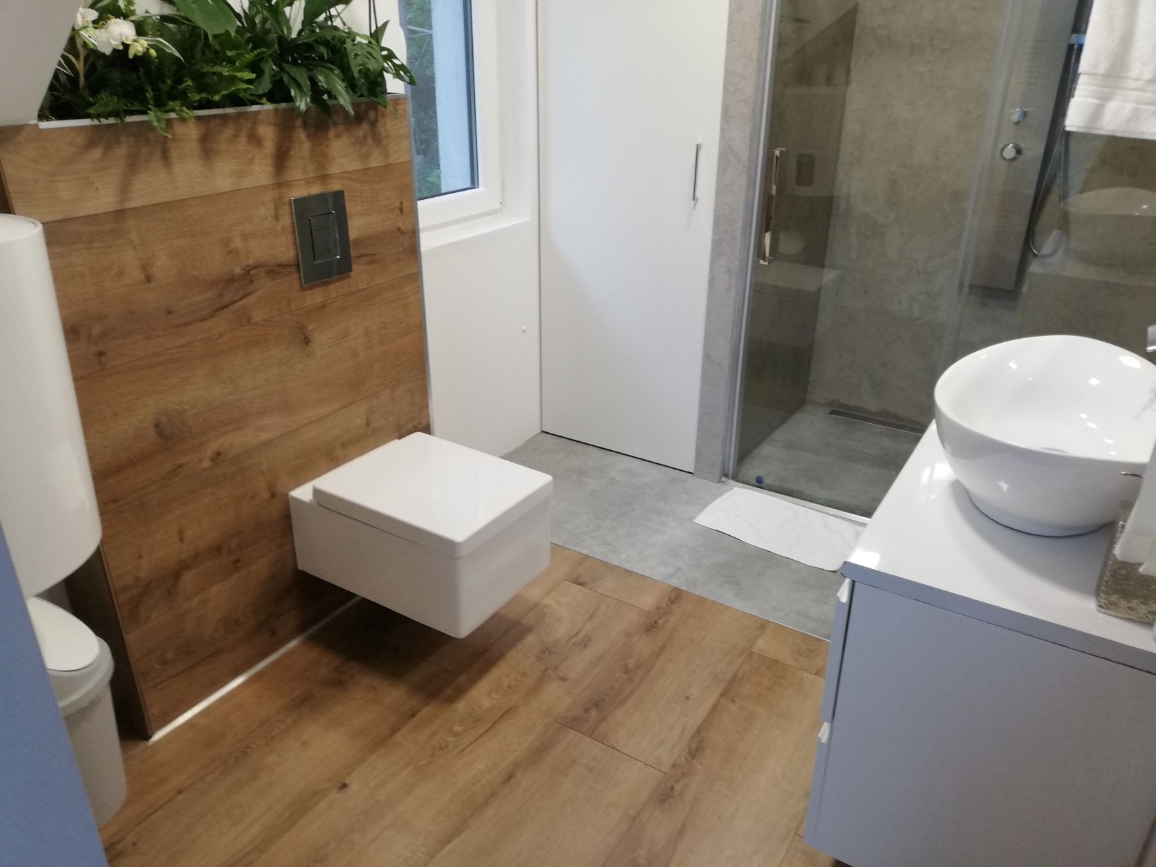 Nowoczesna łazienka z podwieszaną miską WC, drewnianą okładziną ściany z zielonymi roślinami, szklaną kabiną prysznicową i umywalką nablatową.