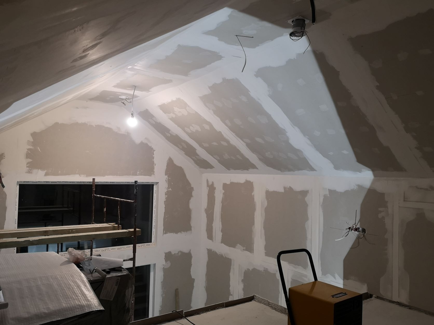 Poddasze w trakcie wykańczania: ściany i skośny sufit pokryte płytami gipsowo-kartonowymi, częściowo wyszpachlowane, widoczne łączenia płyt, prowizoryczne oświetlenie żarówką, rusztowanie...