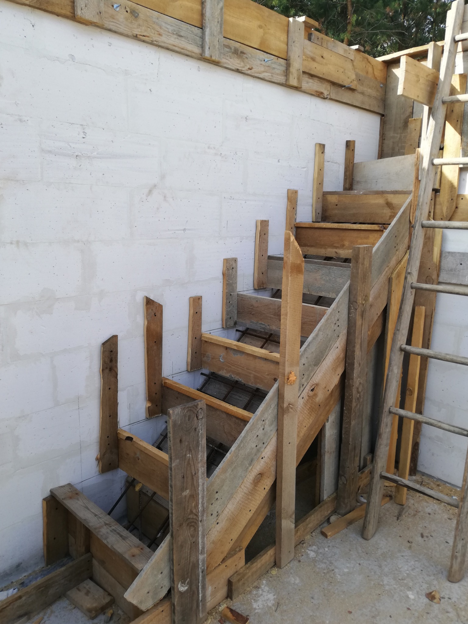 Szare betonowe ściany fundamentowe z drewnianym szalunkiem schodów, widoczna stalowa konstrukcja zbrojeniowa wewnątrz szalunku.