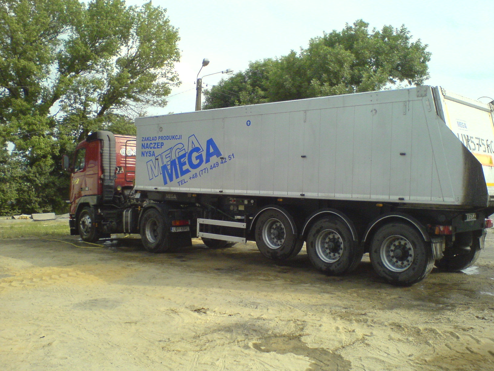 Czerwona ciężarówka z naczepą firmy MEGA z Nysy, zaparkowana na piaszczystym placu.