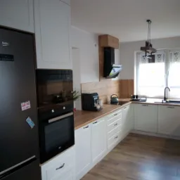 Jasna kuchnia z drewnianymi blatami i czarnym sprzętem AGD, widok na okno z roślinami, na lodówce przyczepione magnesy.