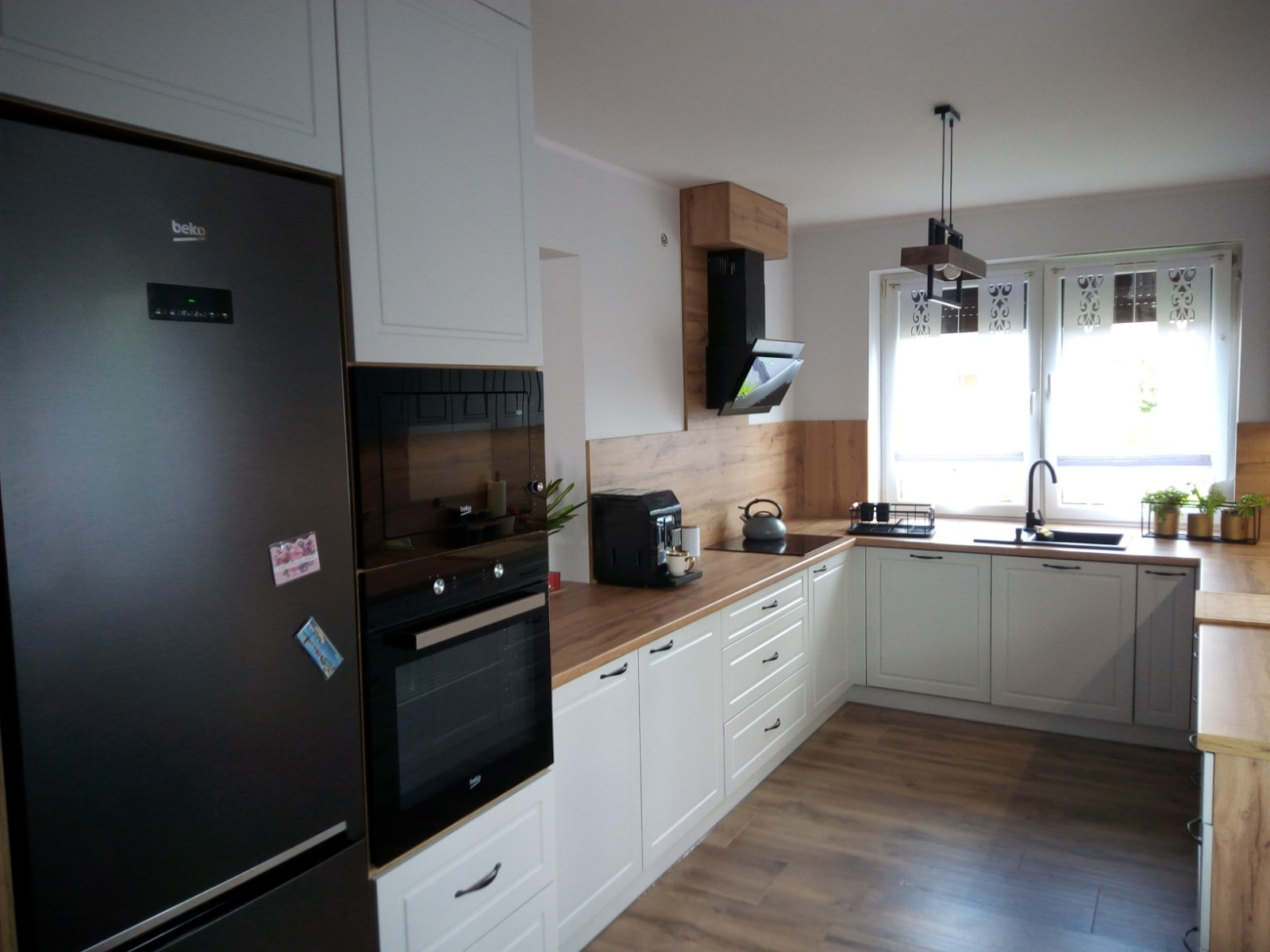 Jasna kuchnia z drewnianymi blatami i czarnym sprzętem AGD, widok na okno z roślinami, na lodówce przyczepione magnesy.