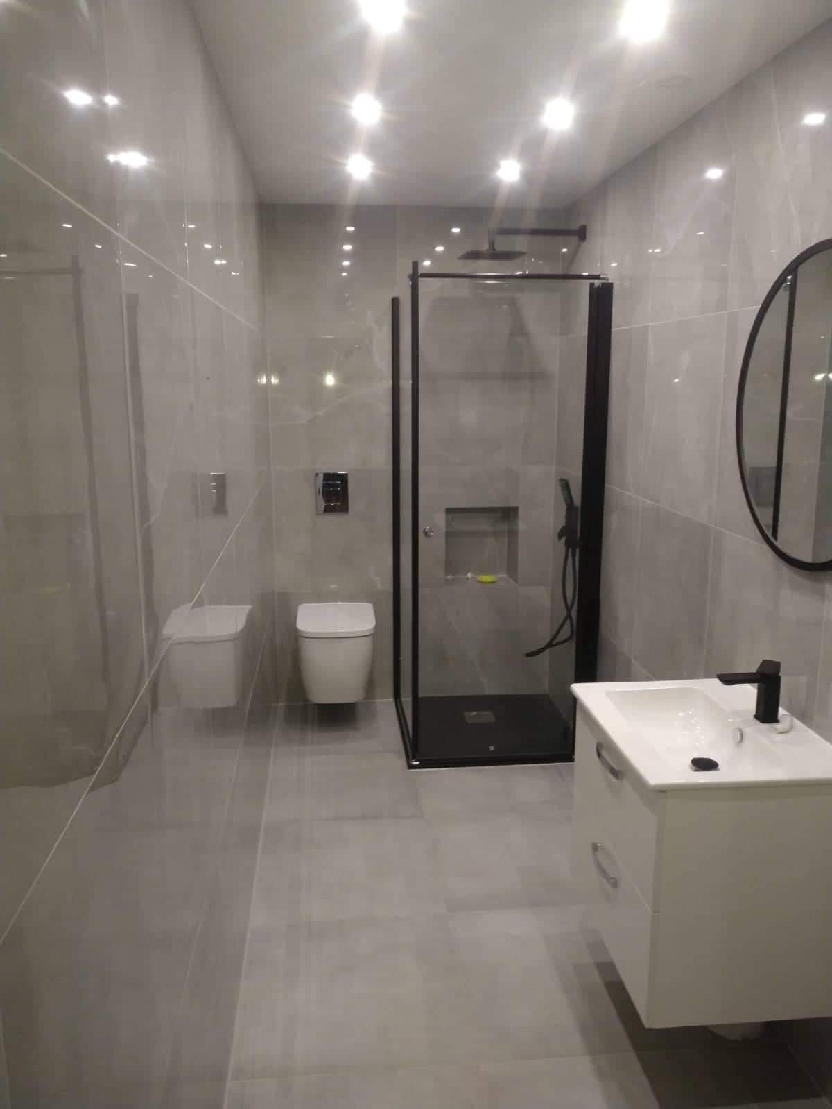 Nowoczesna łazienka z szarymi płytkami, kabiną prysznicową z czarnymi ramami, podwieszaną toaletą i umywalką z szafką w minimalistycznym stylu.