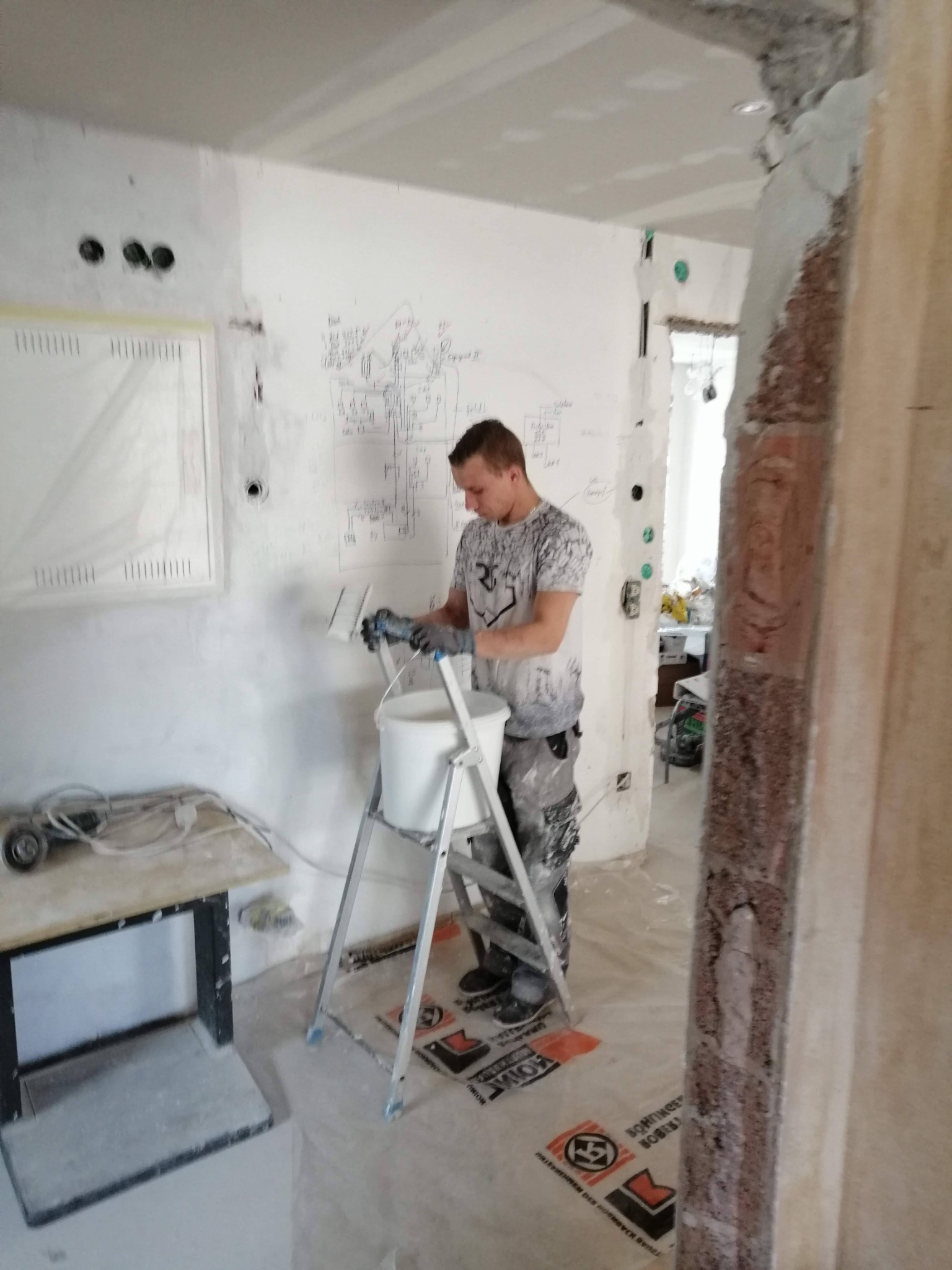 Mężczyzna w rękawicach maluje białą farbą ścianę w pomieszczeniu w trakcie remontu, widoczny schemat instalacji elektrycznej na ścianie, drabina malarska i osłonięta folią futryna drzwi.