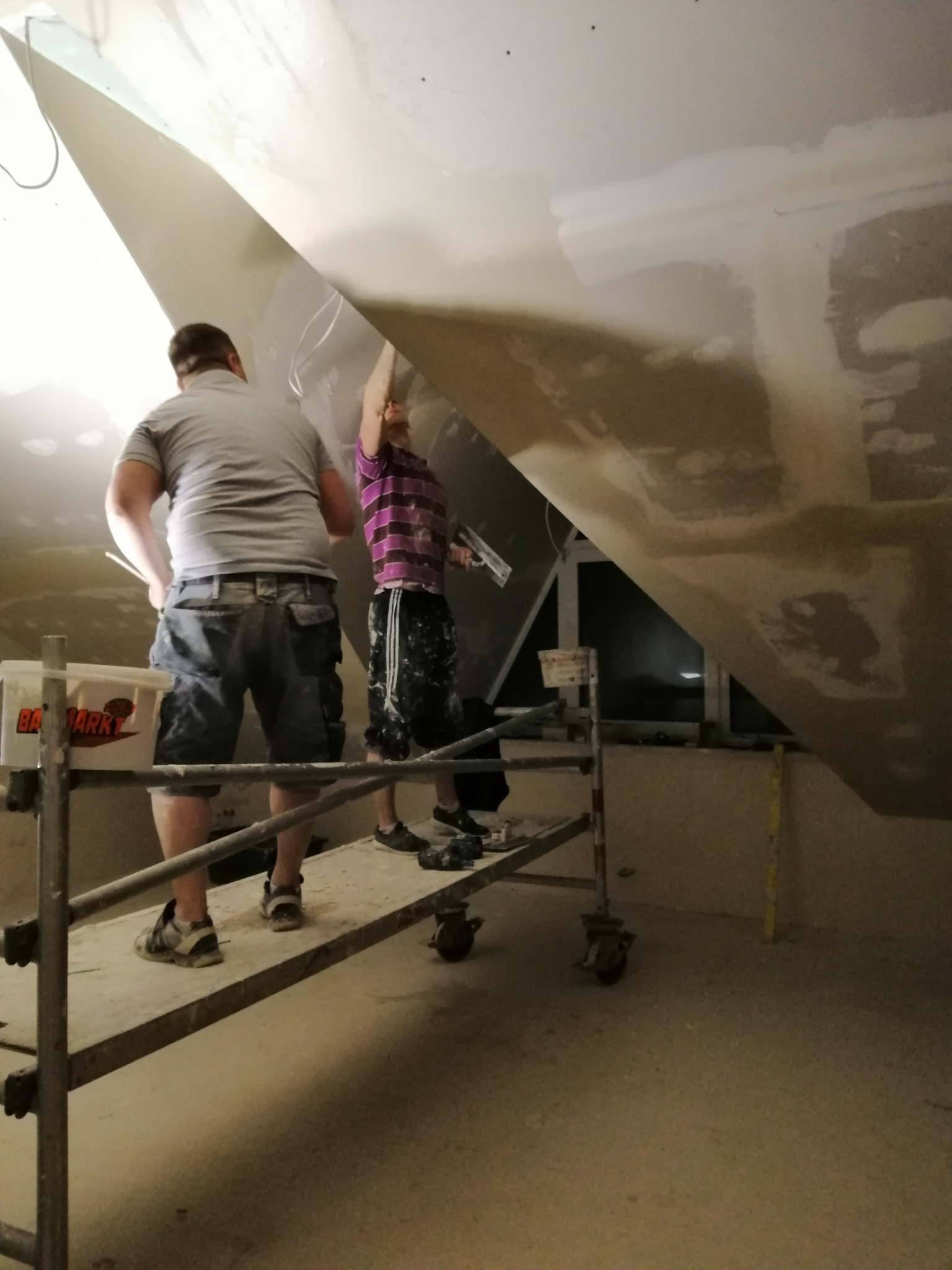 Dwóch mężczyzn na rusztowaniu montuje płytę gipsowo-kartonową na suficie pod skosem w trakcie remontu.