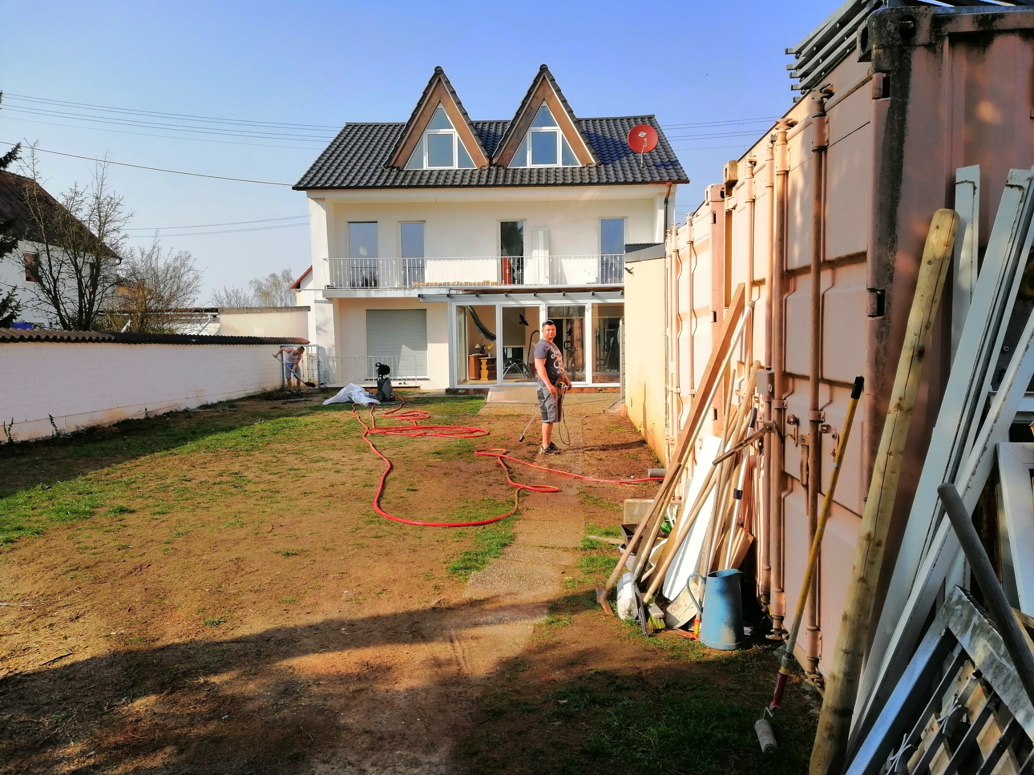 Pracownik myje elewację domu jednorodzinnego z dwuspadowym dachem i oknami trójkątnymi, widoczna czerwona rura i narzędzia budowlane przy kontenerze.