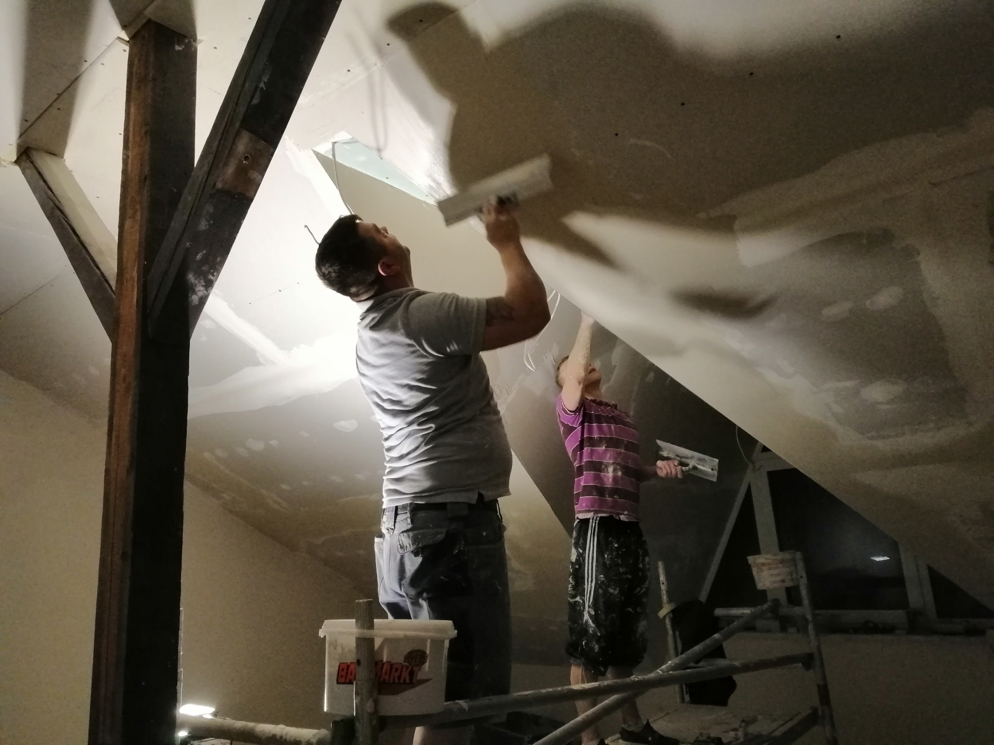 Dwóch mężczyzn wygładza szpachlą sufit poddasza, widoczne drewniane belki konstrukcyjne i pojemnik z masą szpachlową.