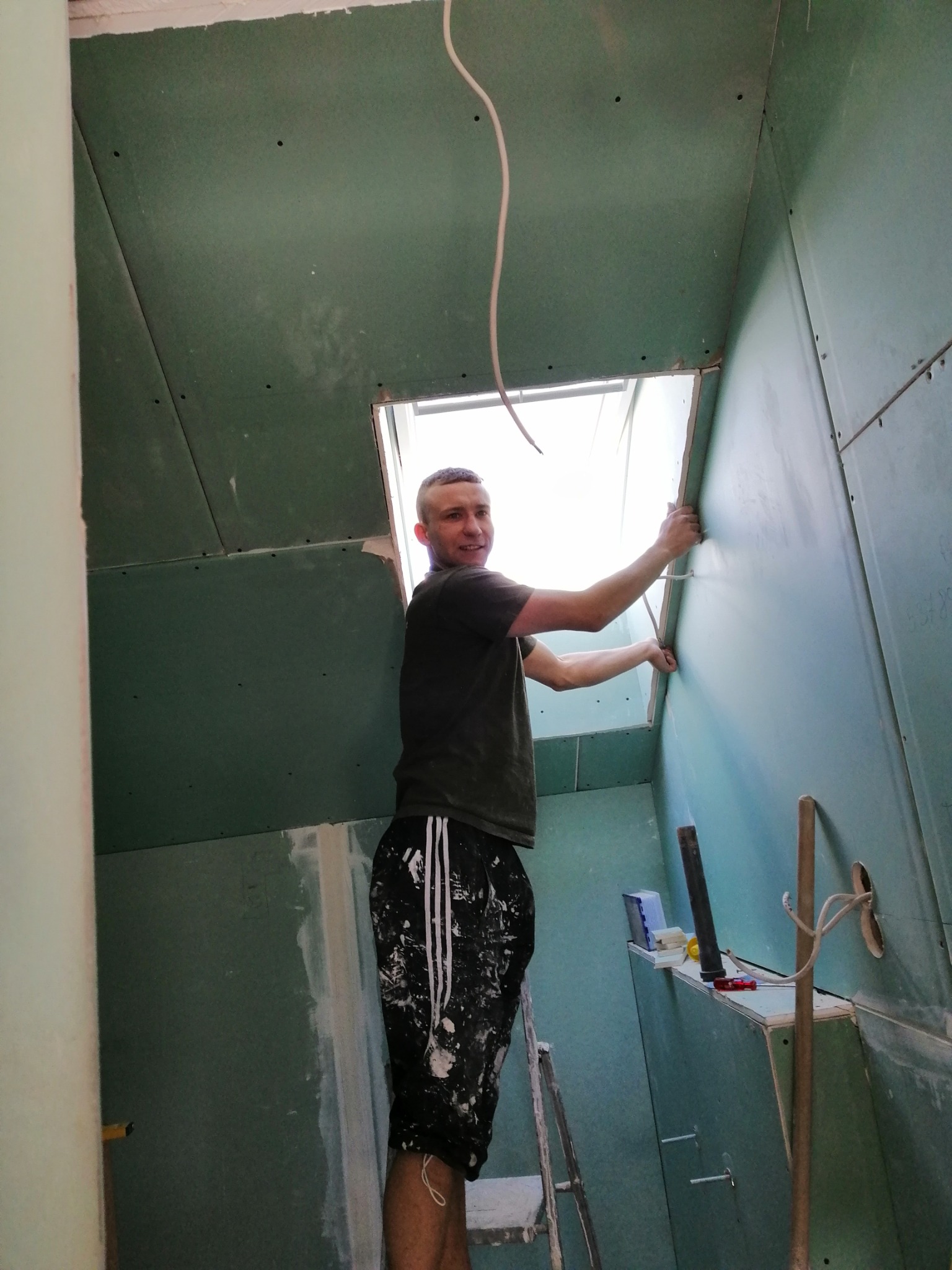 Młody mężczyzna w sportowych spodenkach z plamami gipsu montuje zieloną płytę kartonowo-gipsową w pomieszczeniu, widoczna drabina, narzędzia i wystający kabel.