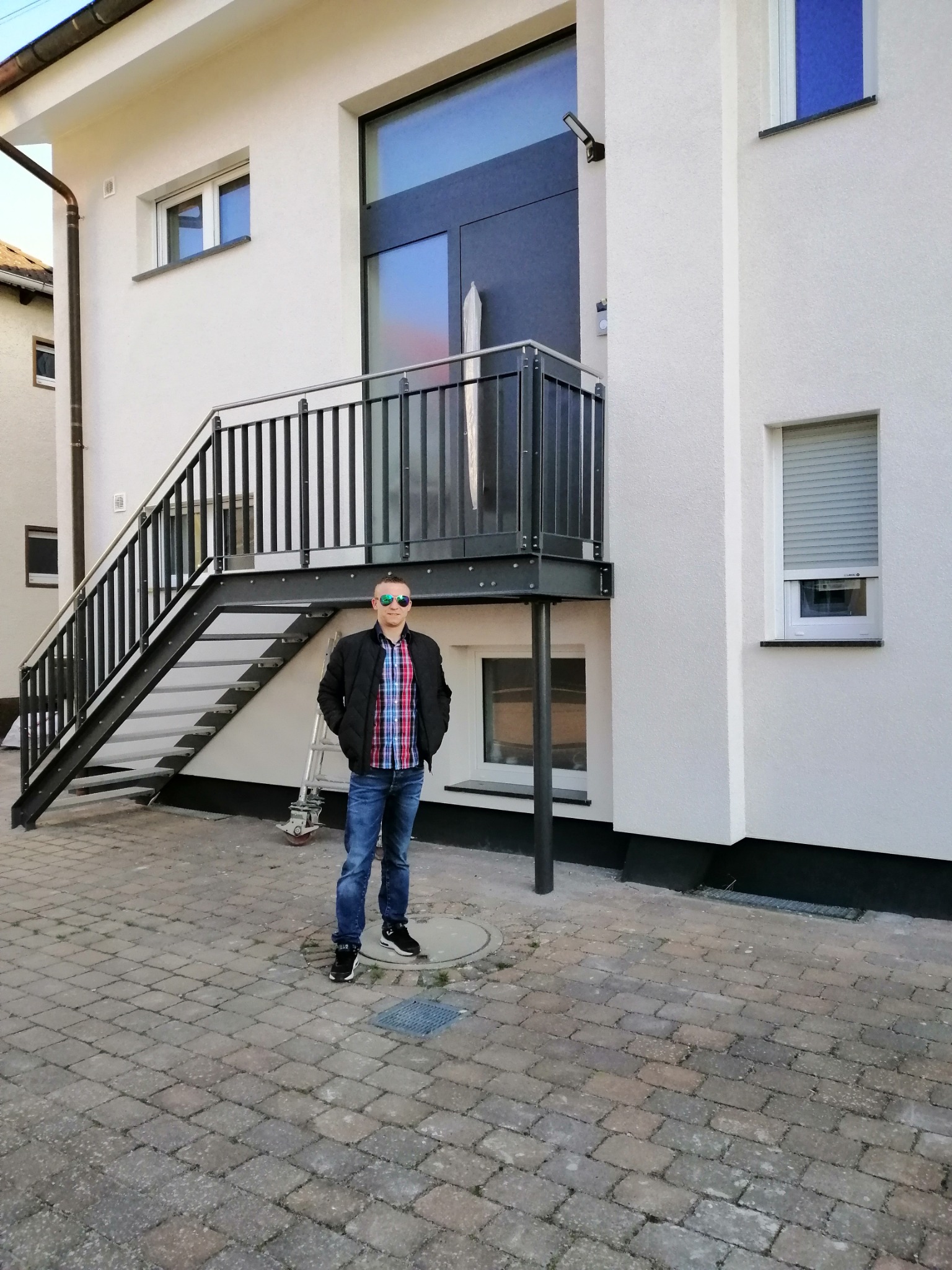 Ocieplony budynek z zewnętrznymi schodami stalowymi, okna z roletami i mężczyzna w okularach przeciwsłonecznych stojący na kostce brukowej.